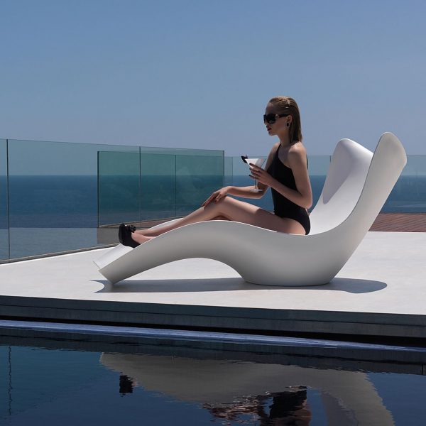 Image of woman sat with drink in white Surf modern reclining chair by Vondom on sunny poolside with glass screen and sea and sky in the background