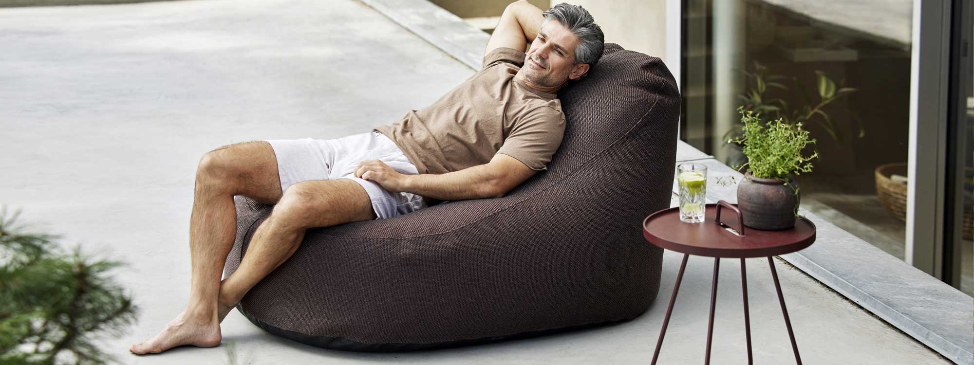 Image of man lying back in Cozy bean bag in Dark Bordeaux fabric, next to On The Move tray table by Cane-line garden furniture