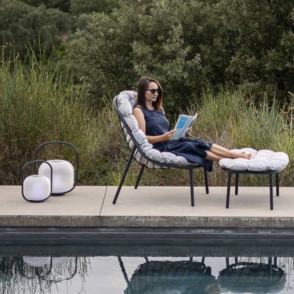 Image of woman sat on Albus lounge chair with her feet resting on foot stool, shown in poolside with Otus outdoor LED lanterns to one side