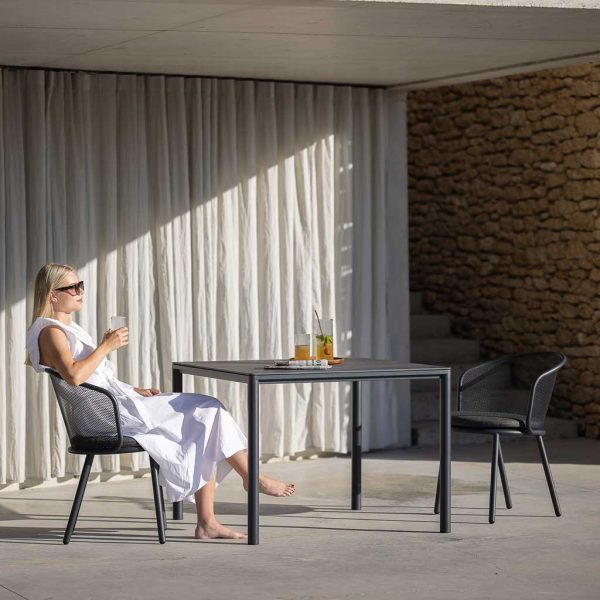Image of woman in sunglasses sat with drink in Baza garden chair next to Alca small garden table in anthracite stainless steel