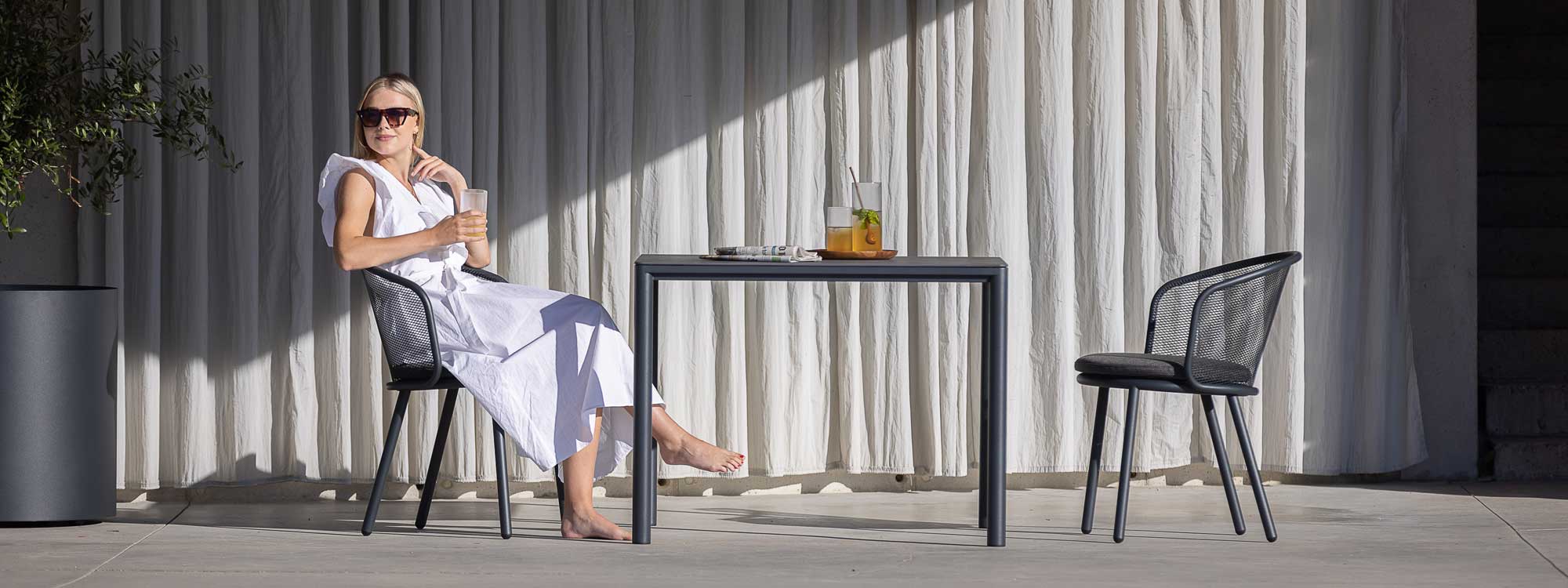 Image of woman sat in Baza garden chair next to Alca square garden table on minimalist poolside terrace