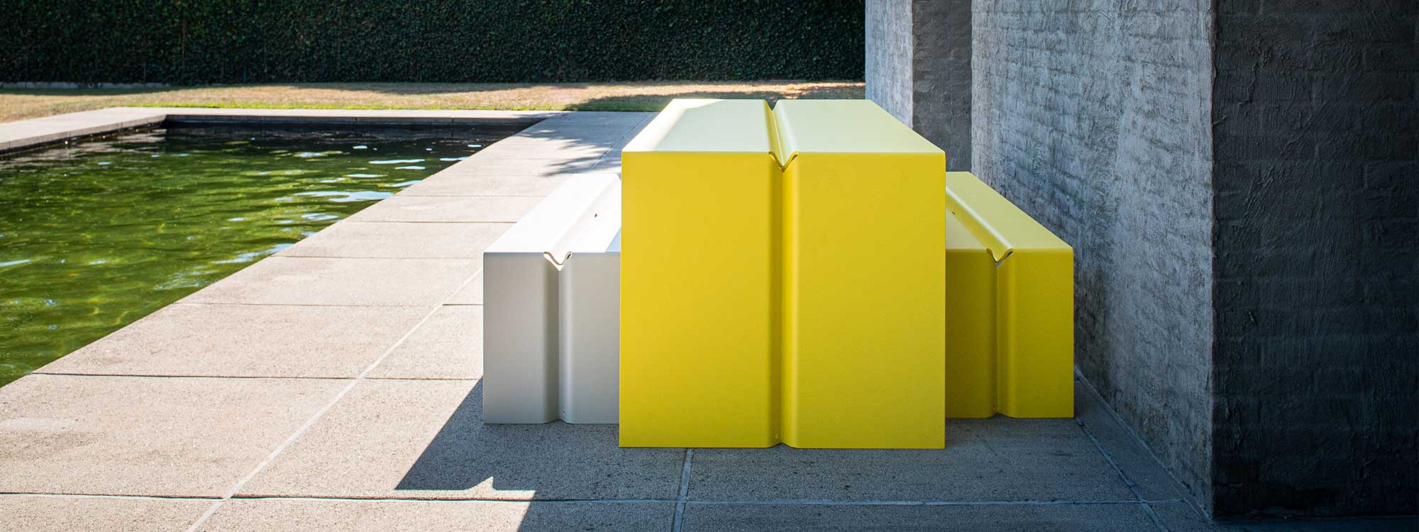 Image of Wünder's The Bended contemporary yellow garden table and white and yellow bench seats in shaped aluminium, shown in courtyard next to peaceful water feature