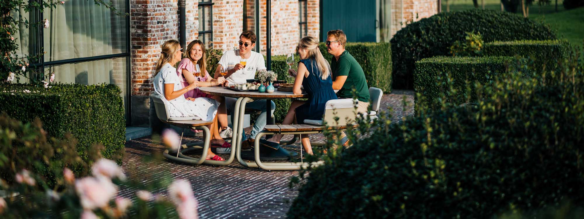 Image of friends sat drinking beers at The Circle round picnic table and benches by Wunder, shown on sunny cobbled floor with old brick building and Buxus shrubs in the background
