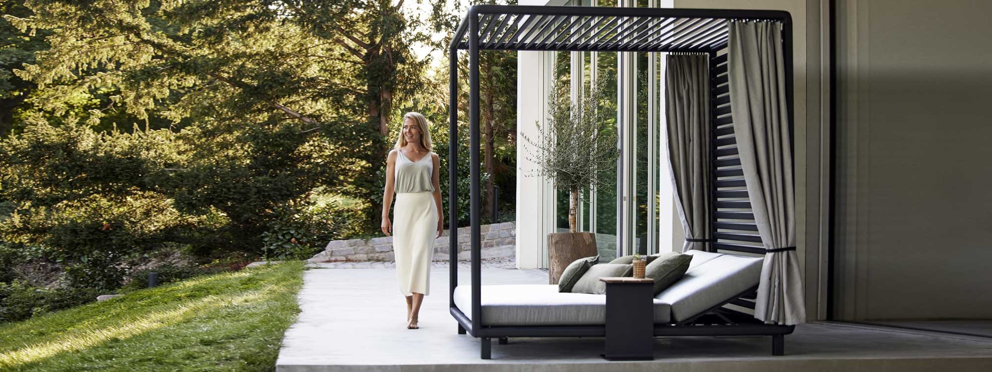 Image of woman walking along terrace next to Laze cabana daybed in lava grey by Cane-line