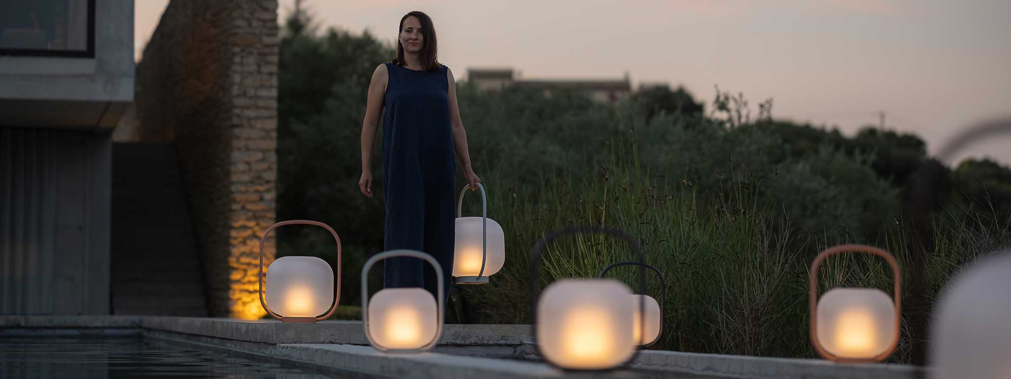 Image at dusk of woman walking along terrace with Otus LED garden lantern, with other lanterns surrounding her on the floor