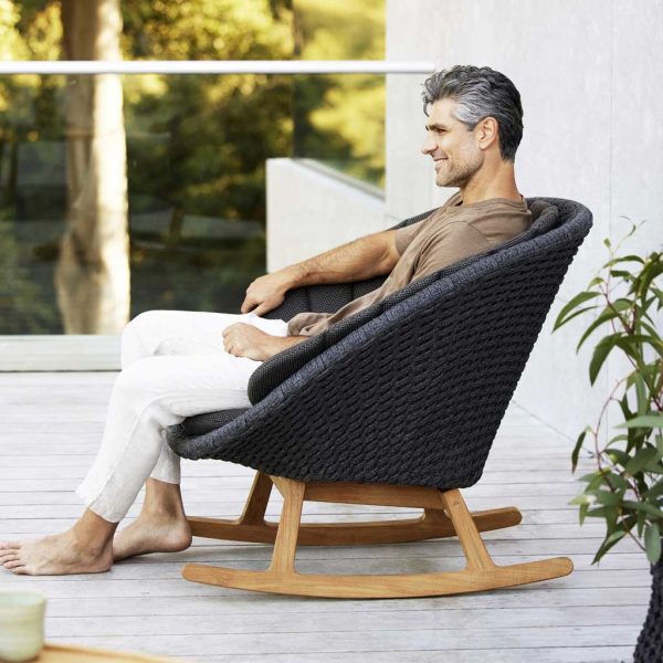 Image of man sat in Peacock garden rocking chair in grey Soft Rope with teak sled legs by Cane-line garden furniture, Denmark.