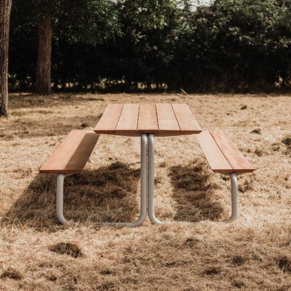 Image of Wünder The Table modern picnic furniture with white tubular steel frame and afzelia hardwood surfaces, shown on scorched grass with woodland in the background