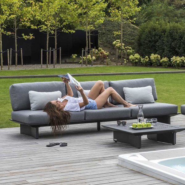 Image of woman lying back reading a magazine on Arbon dark grey garden sofa with grey cushions, shown on sunny poolside with lawn and woodland in the background
