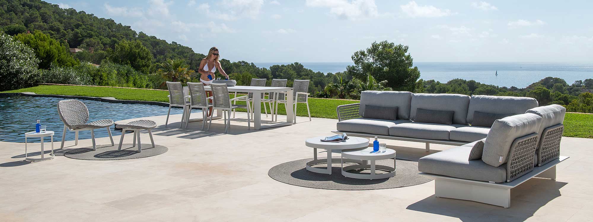 Image of Arbon white garden corner sofa, together with Vigo XL extending table and Sevilla chairs on sunny poolside with lawn, woodland and blue sea and sky in the background