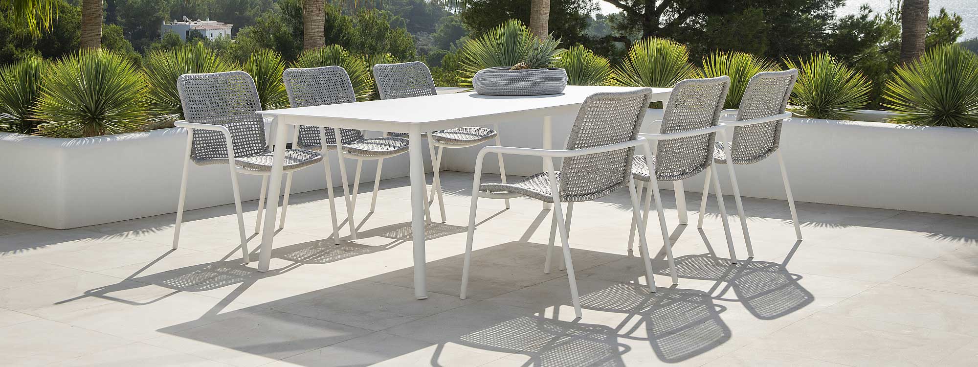 Image of Durham modern garden furniture with white tubular aluminium frames and white-grey Polyolefin rope seats and backs by Jati & Kebon, shown on sunny whitewashed terrace, with exotic plants and the sea in the background