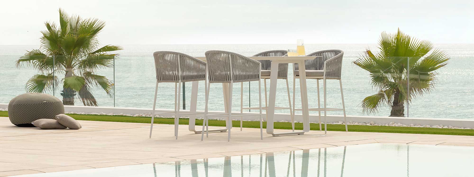 Image of Fortuna Rope outdoor tub bar stools around a Jati & Kebon bar table, shown on terrace with the sea in the background