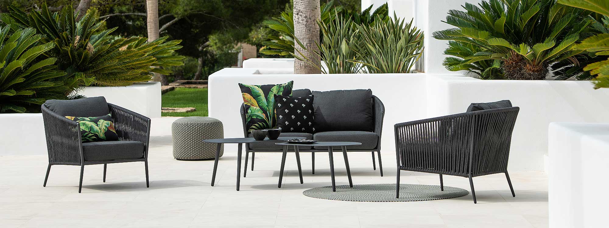 Image of Fortuna Rope outdoor lounge furniture in charcoal black, shown on white-washed terrace surrounded by small palms and bird of paradise plants