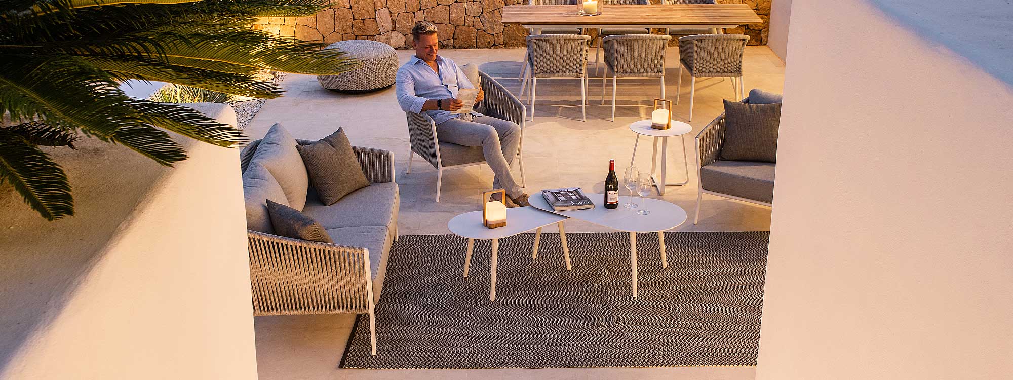 Image of man sat on Fortuna Rope white garden lounge chair with grey-melange woven back, shown on illuminated white-washed terrace at night