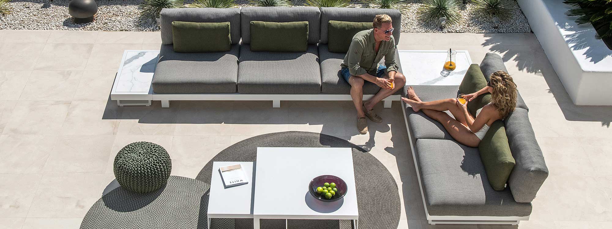Image of aerial view of couple chatting on Fano contemporary garden sofa in white, with grey cushions and side tables with graduario ceramic table tops