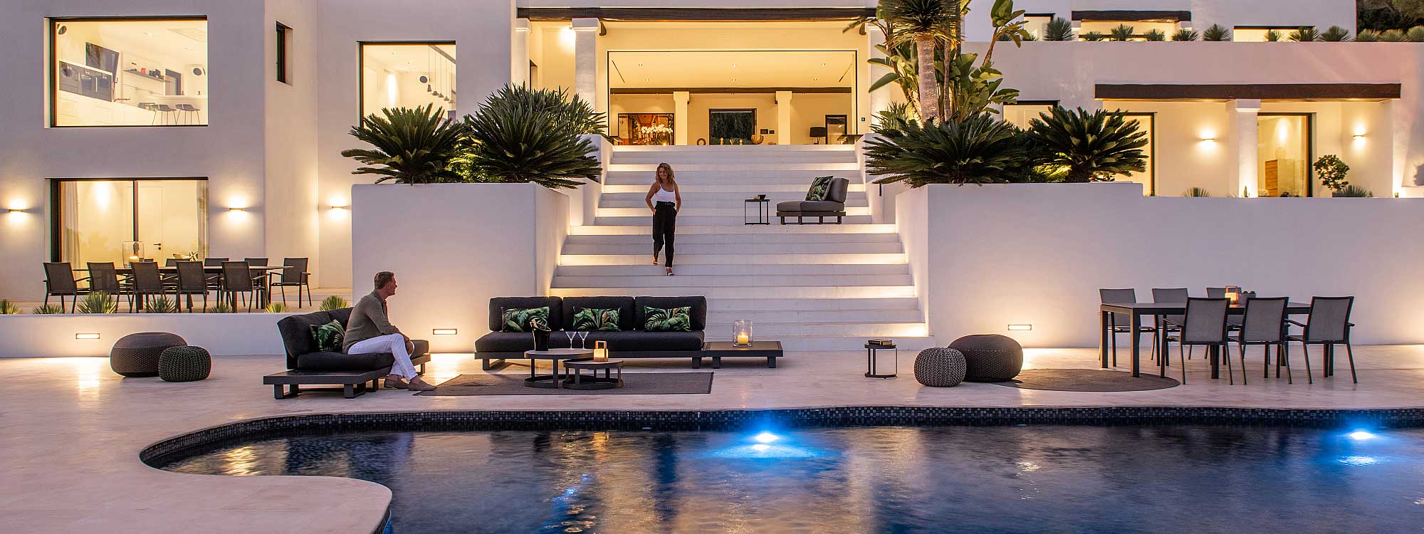 Image of large illuminated night time poolside with man sat on Fano grey garden sofa as woman descends stairs from the house above.