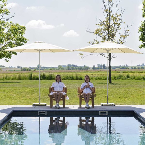 Image of couple of girls sat on poolside beneath pair of small mast parasols by Prostor with Belgian countryside in the background