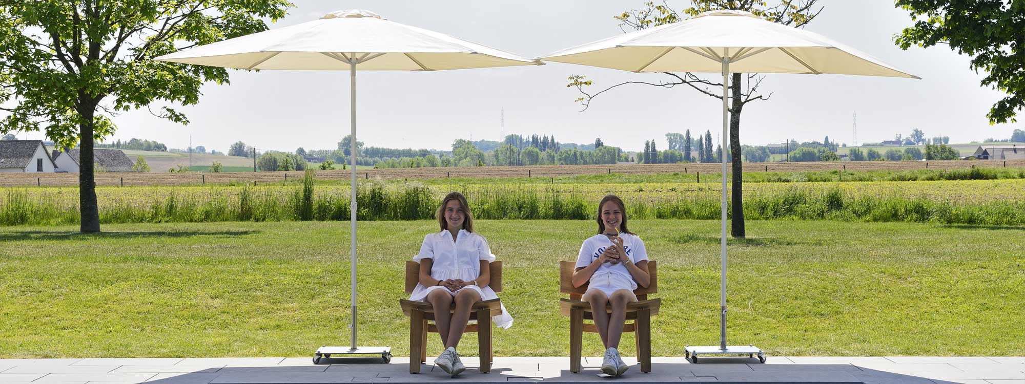Image of 2 girls sat beneath pair of P50 white centre pole parasols by Prostor, with Belgian countryside in the background