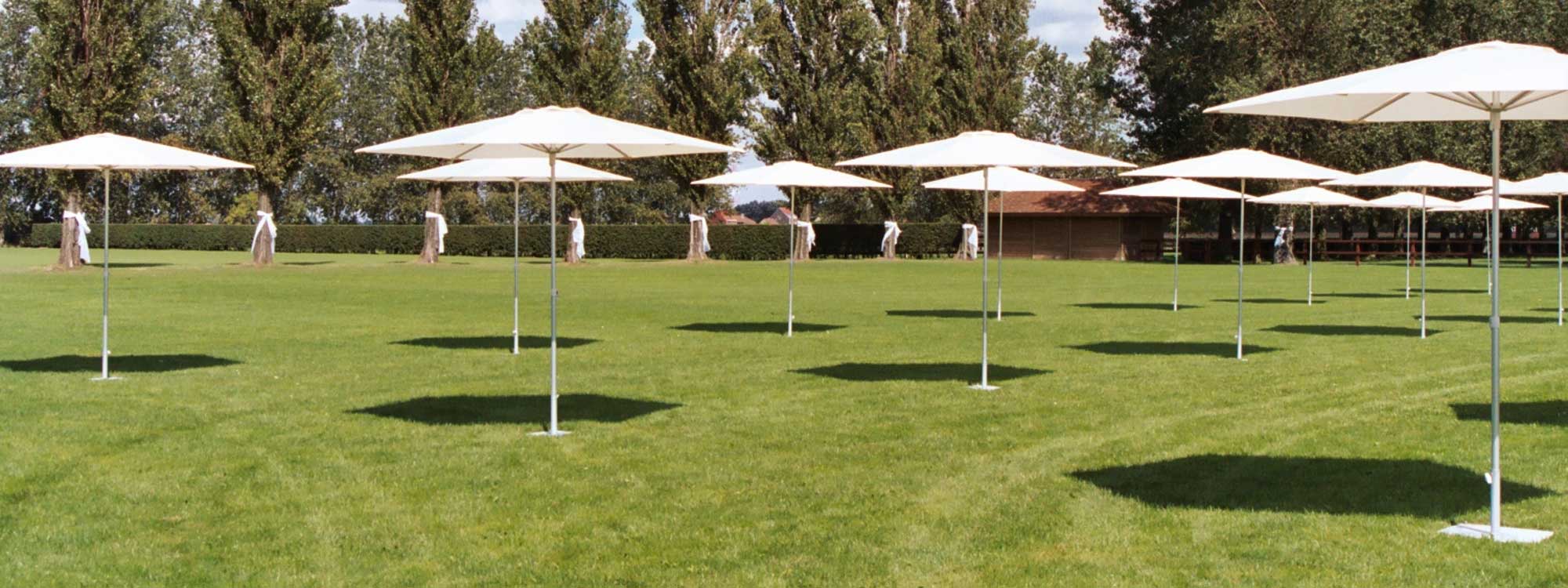 Image of multiple Prostor P50 white mast parasols on a grass lawn, with trees and sky in the background