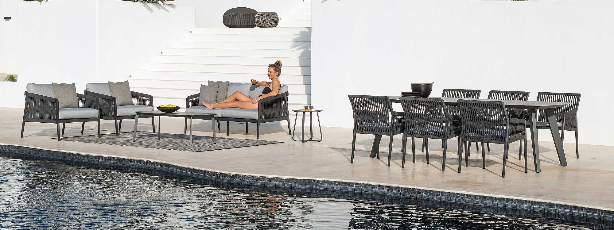 Image of woman relaxing on Ritz black garden sofa on poolside, next to Ritz ceramic garden table and chairs