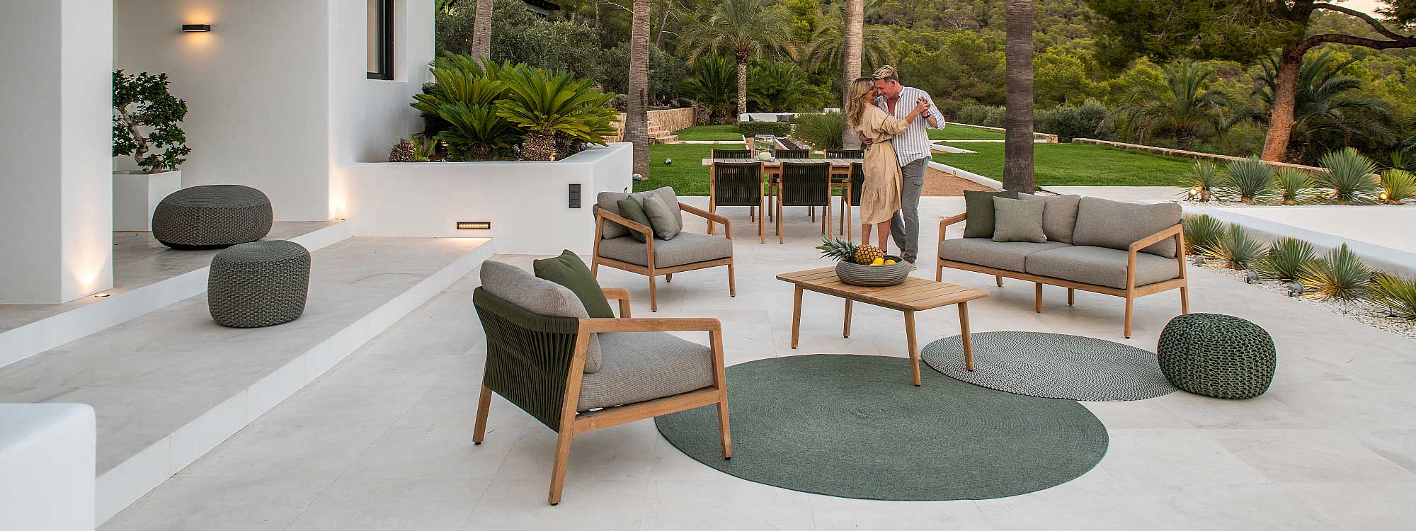 Image of couple dance on expansive terrace at dusk, amongst Ritz teak garden furniture, with lawn and exotic planting in the background