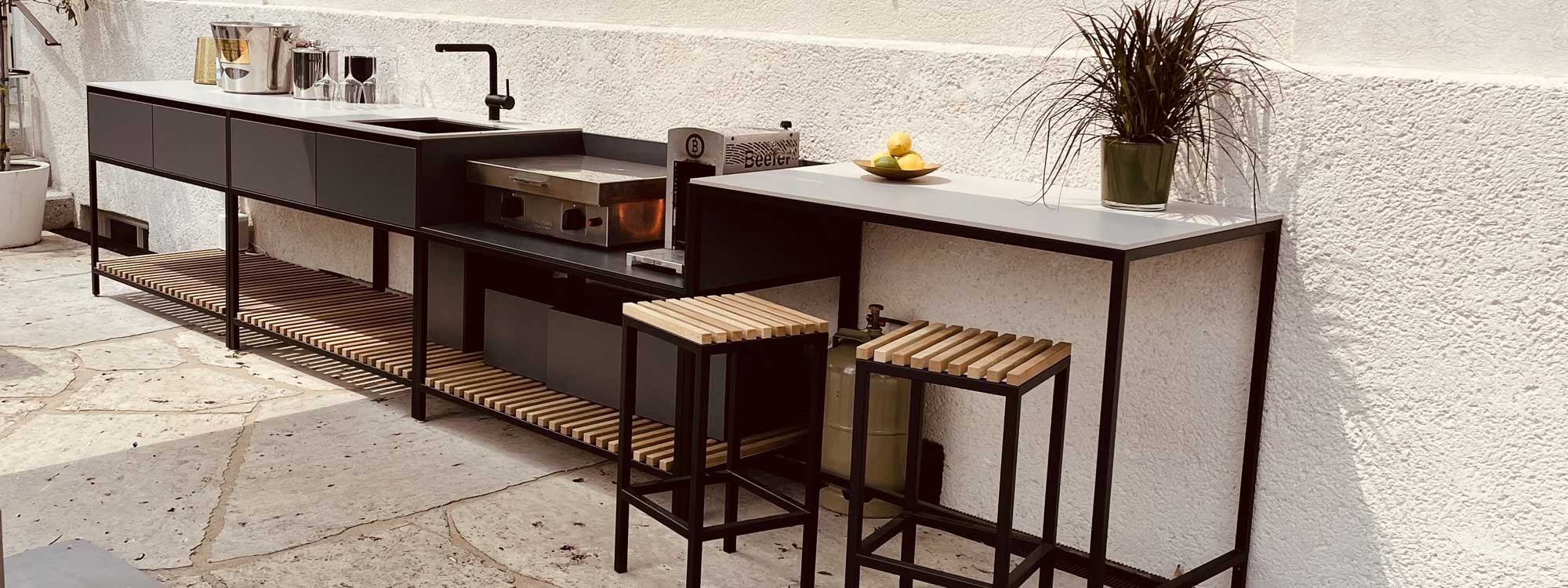 Image of Conmoto Ticino Frame modern garden kitchen with anthracite stainless steel frame and light grey ceramic work tops, shown against whitewashed building