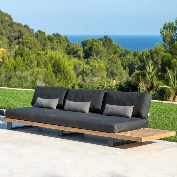 Image of Truro Lounge modular teak garden sofa with dark grey cushions on terrace, with lawn, woodland and sea in the background