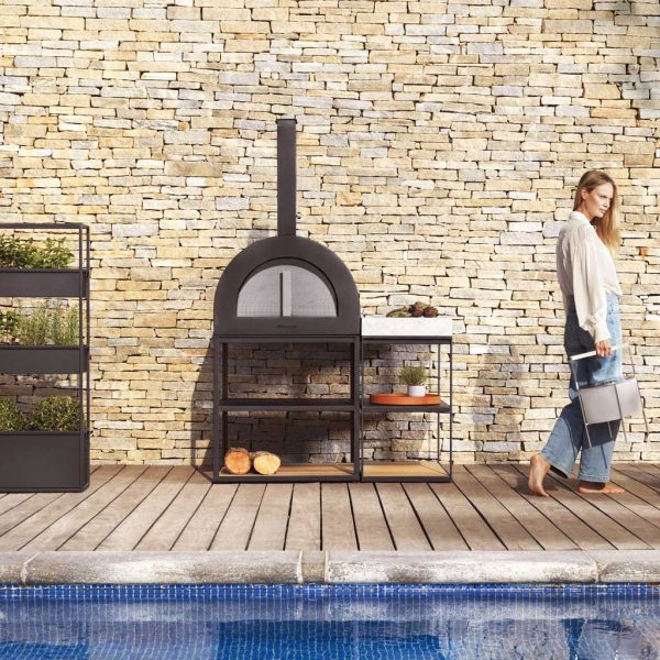 Image of woman on wooden decked poolside walking past Roshults wood fired pizza oven in anthracite stainless steel
