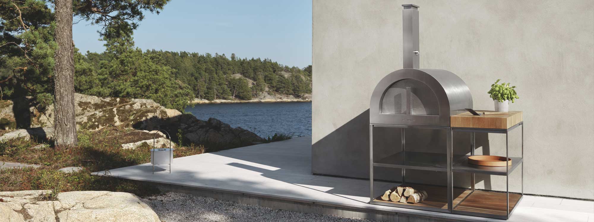 Image of Roshults wood fired pizza oven and outdoor kitchen work top in brushed stainless steel with surfaces in FSC certified teak, shown against side of building, with Swedish lake and woodland in the background