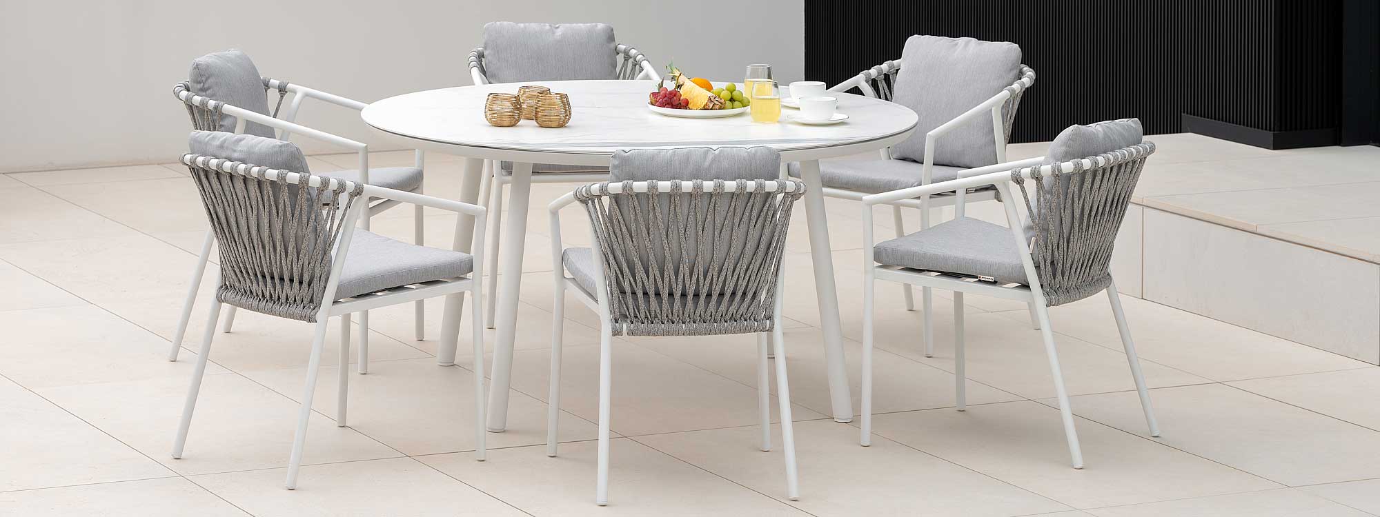 Image of white Kapra garden chairs around Durham round garden dining table, with bowl of fruit and drinks placed on the table top