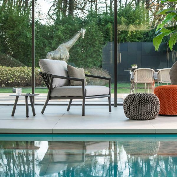 Image of Kapra black outdoor easy chair with light grey cushions, on indoor poolside with floor to ceiling glass in the background