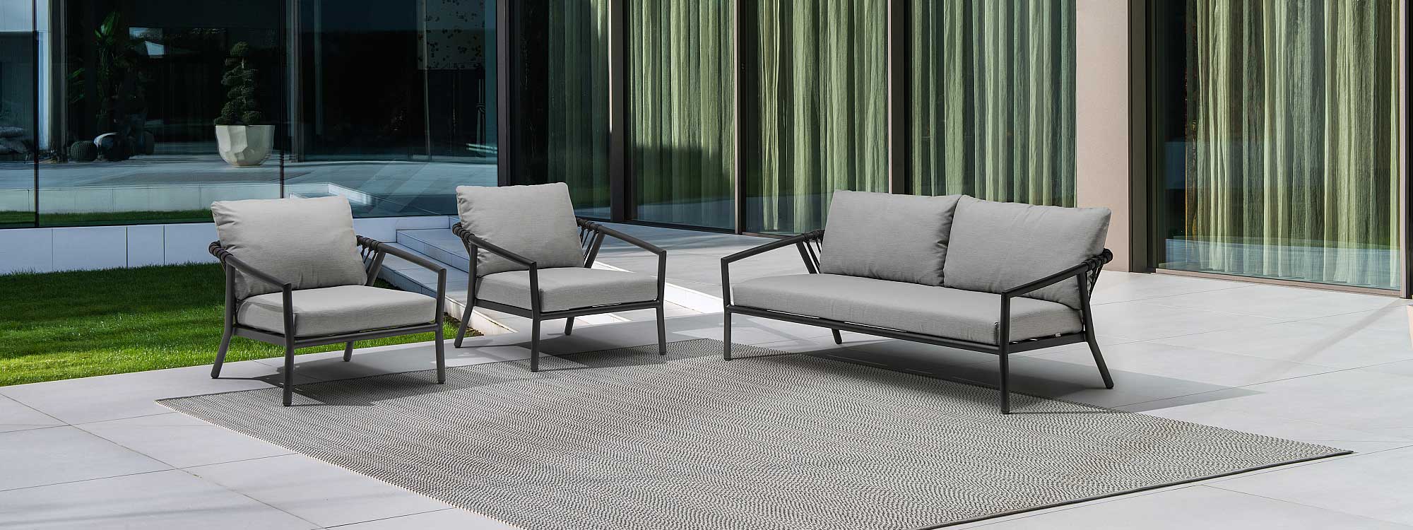 Image of Kapra modern garden sofa and lounge chairs with Charcoal tubular aluminium frames and Charcoal Black woven rope backs on minimalist terrace with floor to ceiling glass windows in the background