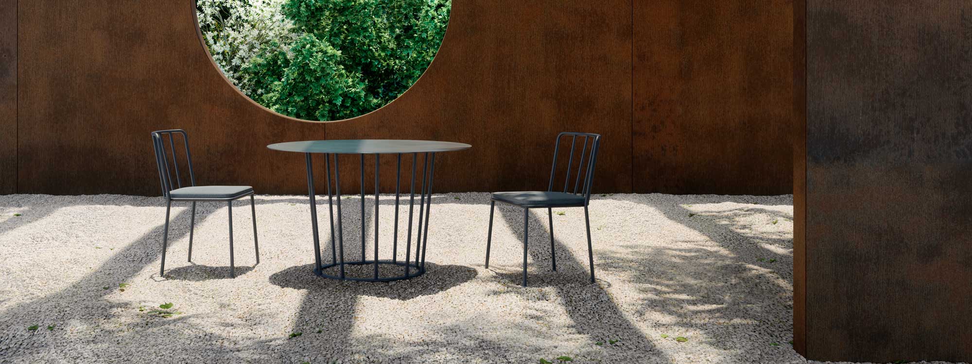 Image of Oiside Panama circular garden table & modern outdoor chairs, on gravel floor with oxidised corten steel structure in the background with round window looking out onto shrubs and trees