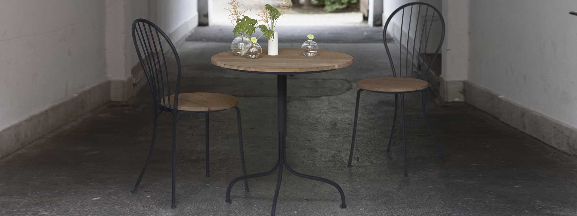 Image of pair of Akleja chairs either side of Akleja table in dark-grey steel and teak by Grythyttan Stålmöbler, shown in outdoor passageway
