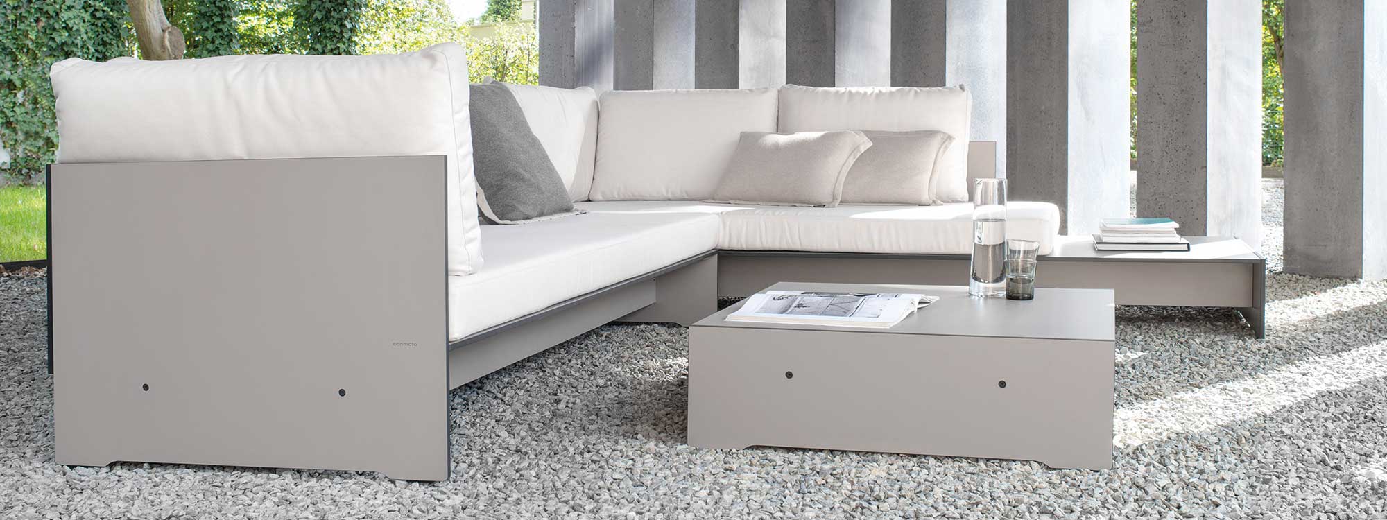 Image of Conmoto Riva modern garden corner sofa in taupe HPL, shown on gravel floor beneath geometric concrete structure