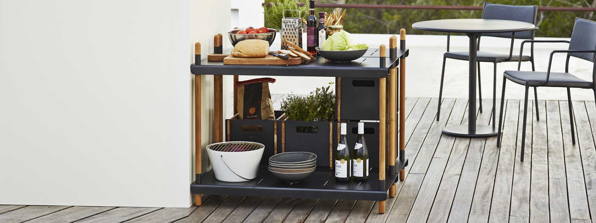 Image of pair of Frame outdoor shelving units in Lava-Grey aluminium and teak, with Go table and Less outdoor chairs by Cane-line in the background