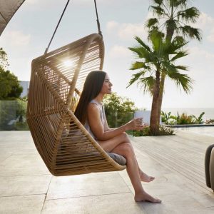Image of woman sat in Cane-line Hive contemporary garden swing seat on balmy seaside terrace at sunset