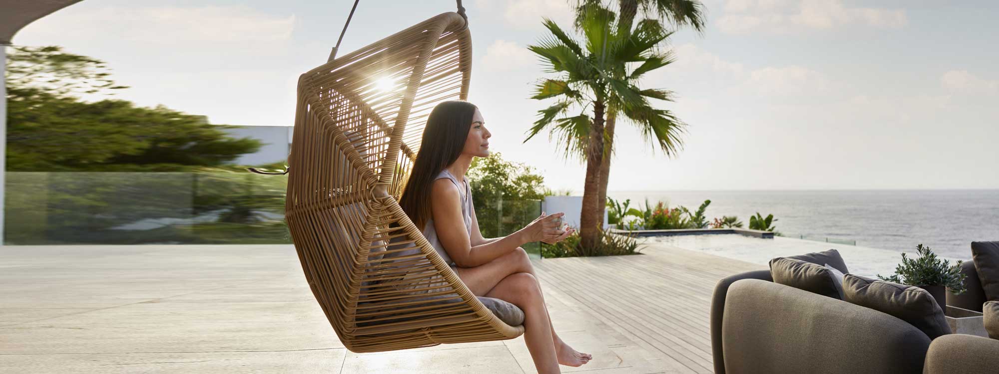 Image of woman sat contemplating in Hive modern garden swing chair by Cane-line on seaside terrace at sunset