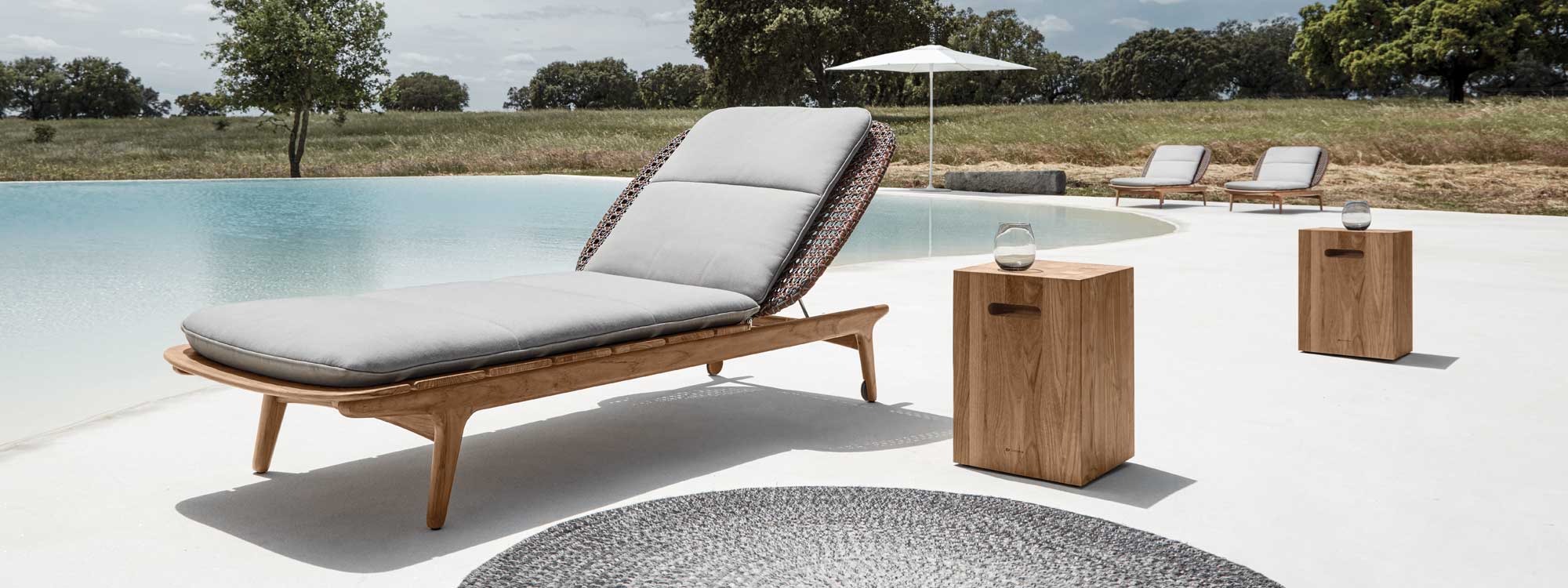 Image of Gloster Kay teak & wicker sunbeds together with Block side tables around a large swimming pool