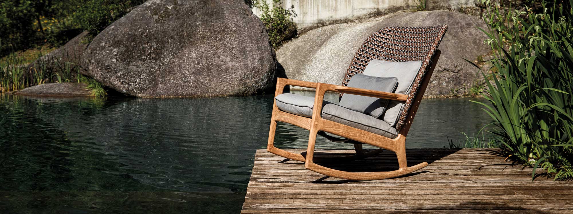 Image of Gloster Kay wicker garden rocking chair on wooden deck next to lake with large boulders in the background