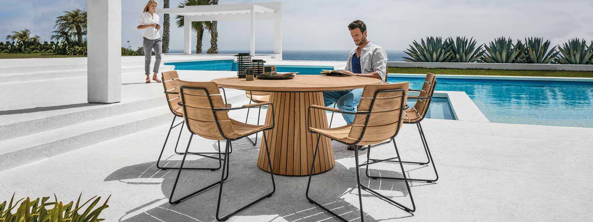 Image of man sat on William teak garden chair next to Whirl unique round table by Gloster