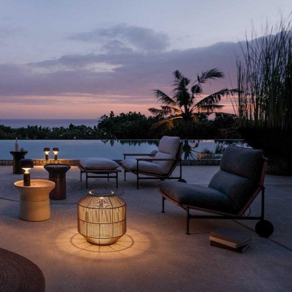 Nighttime image of Gloster Basket garden lantern casting light and shade on terrace, next to Coso side tables and Zenith modern relax furniture
