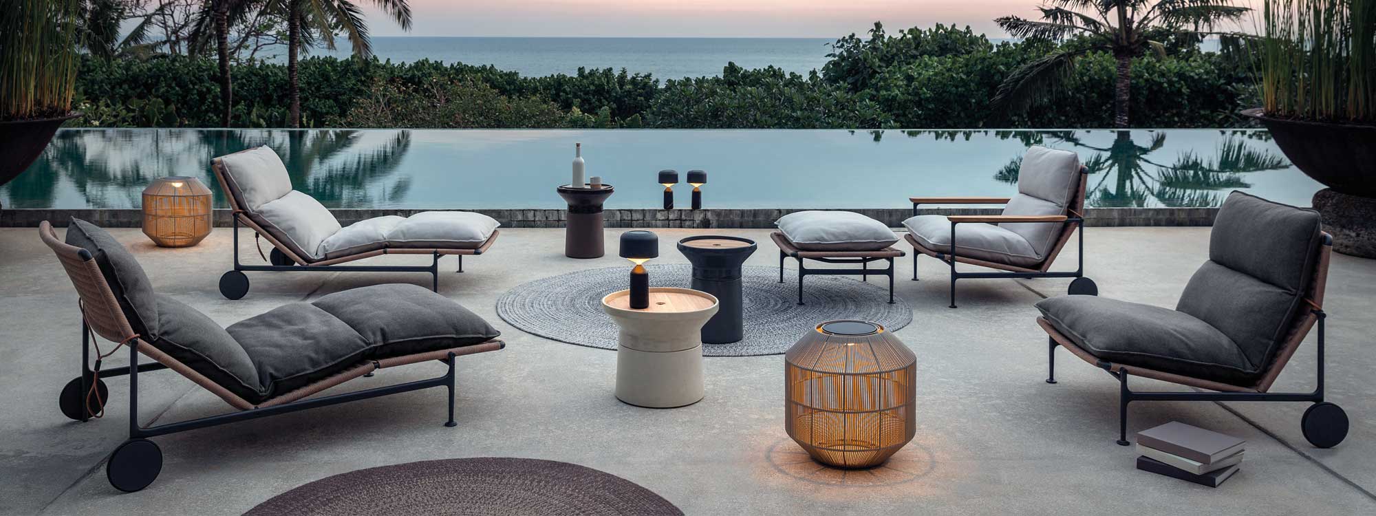Image of Gloster Zenith modern outdoor lounge furniture and illuminated Basket lanterns around peaceful horizon swimming pool at dusk, with tropical woodland and sea in the background