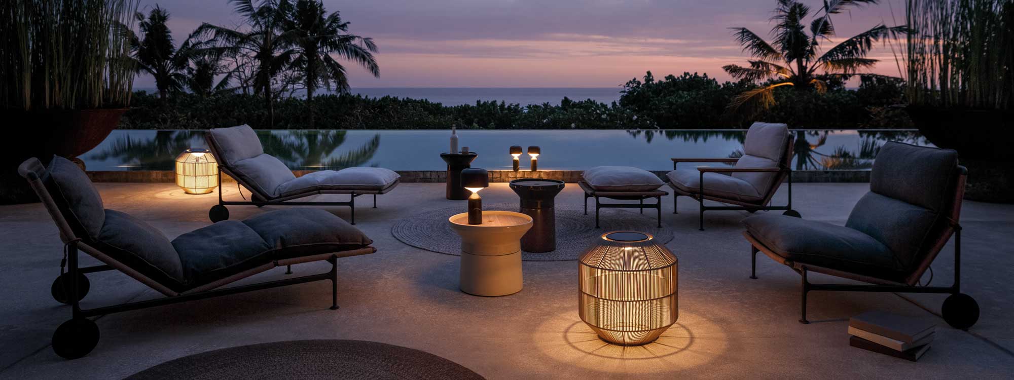 Nighttime image of Gloster Zenhit outdoor lounge furniture, illuminated Basket lanterns and Coso side tables alongside peaceful horizon swimming pool