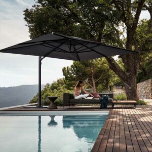 Image of terraced poolside with woman sat reading on Grid sofa in the shade of Gloster Halo side post parasol