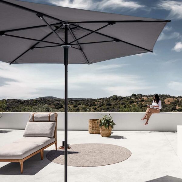 Image of Gloster Halo grey garden parasol with teak chaise longue in the background, together with a woman sat reading on a white-washed wall in the sunshine