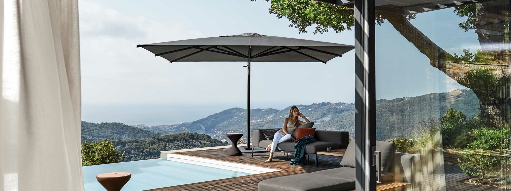 Image of woman sat on Grid sectional garden sofa beneath Halo luxury garden parasol by Gloster, with undulating countryside and distant coastline in the background