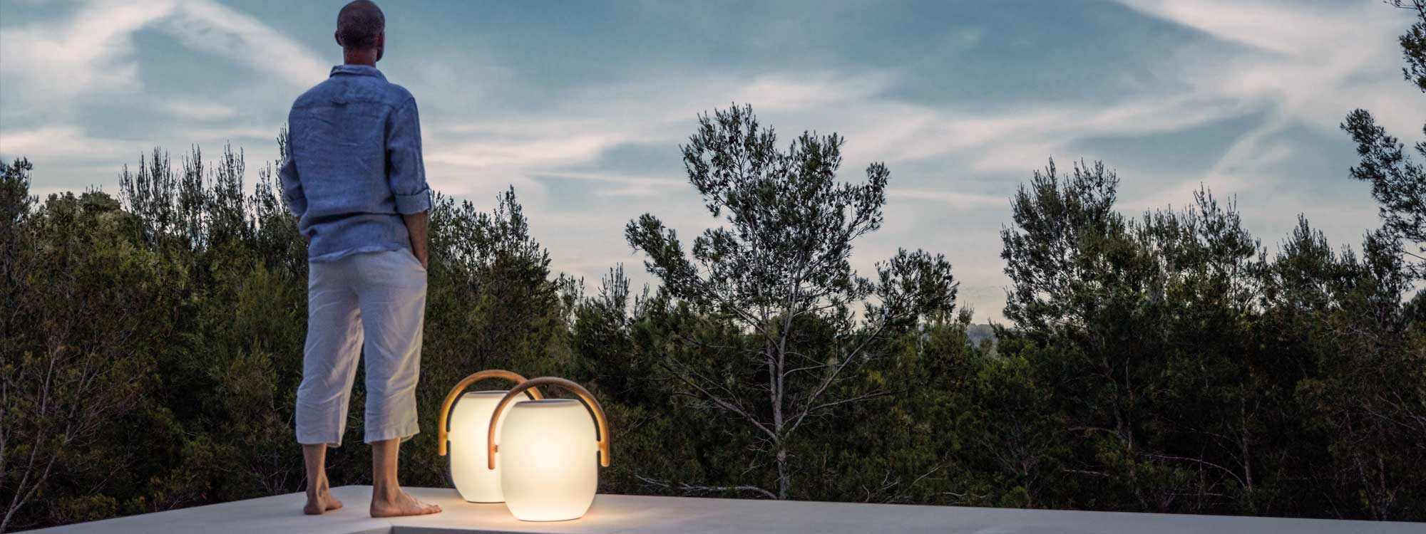 Image of man stood on Mediterranean terrace at dusk next to pair of Gloster Ambient Cocoon modern garden lanterns