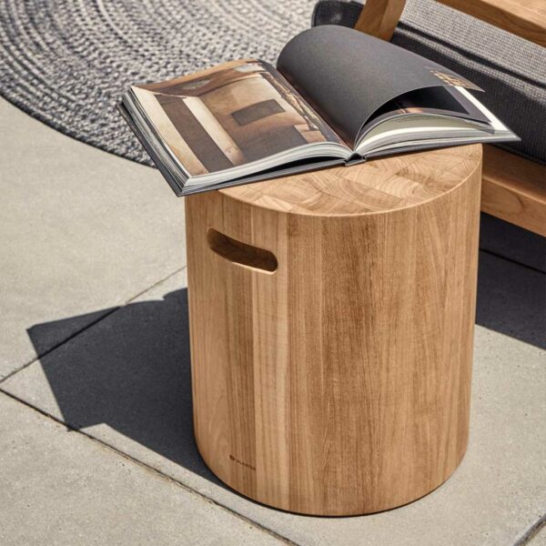 Image of cylindrical Block outdoor teak side table by Gloster with open magazine on the table top