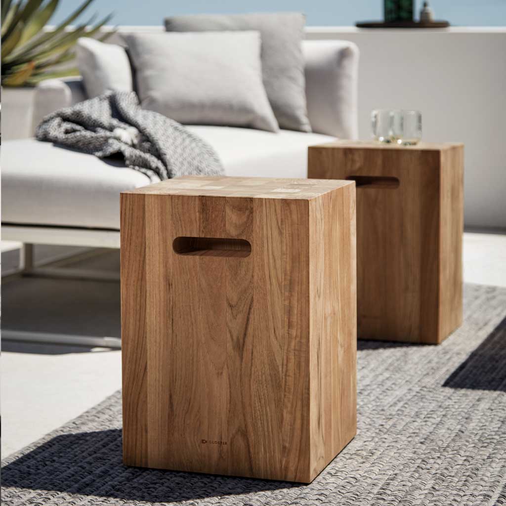 Image of pair of Gloster Block square solid teak side tables in the sun, with Gloster Maya garden sofa in the background