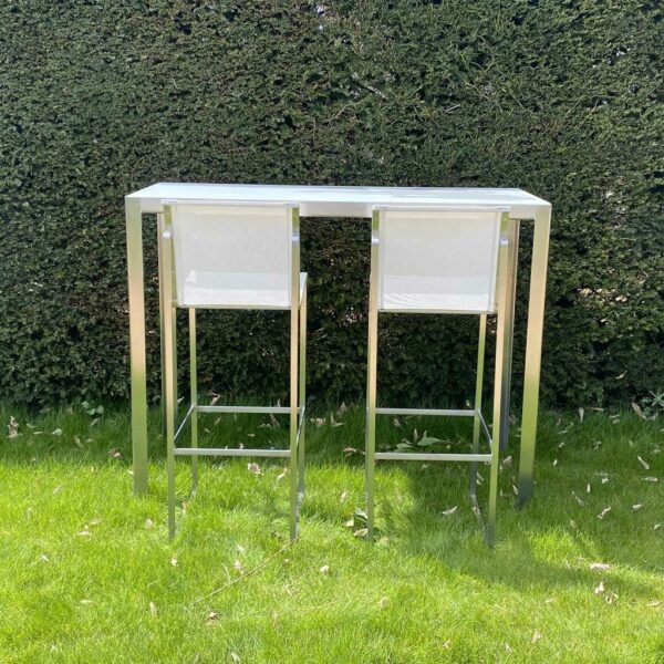 Image of FueraDentro Nimio exterior bar table and Taburete bar stools on lawn against a clipped yew hedge.